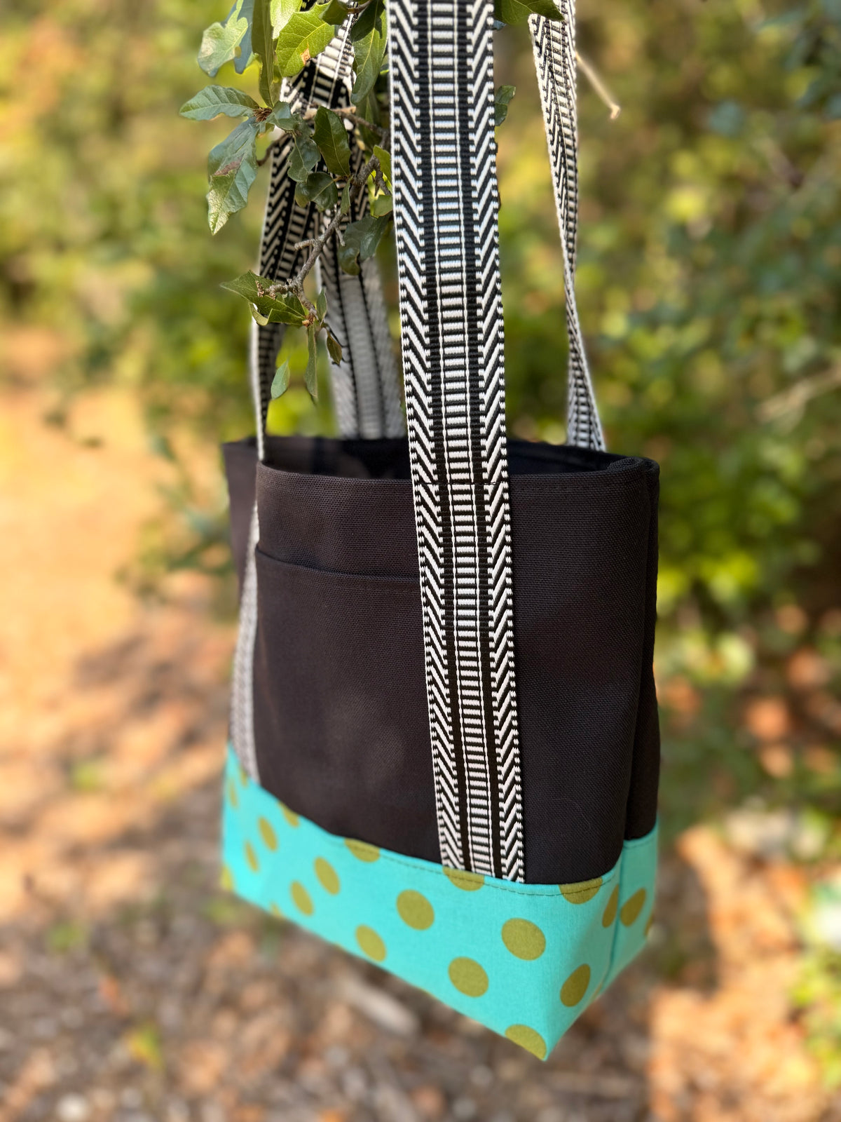 A fabric tote bag  featuring a black upper section, black-and-white patterned handles, and a turquoise bottom panel with green polka dots.