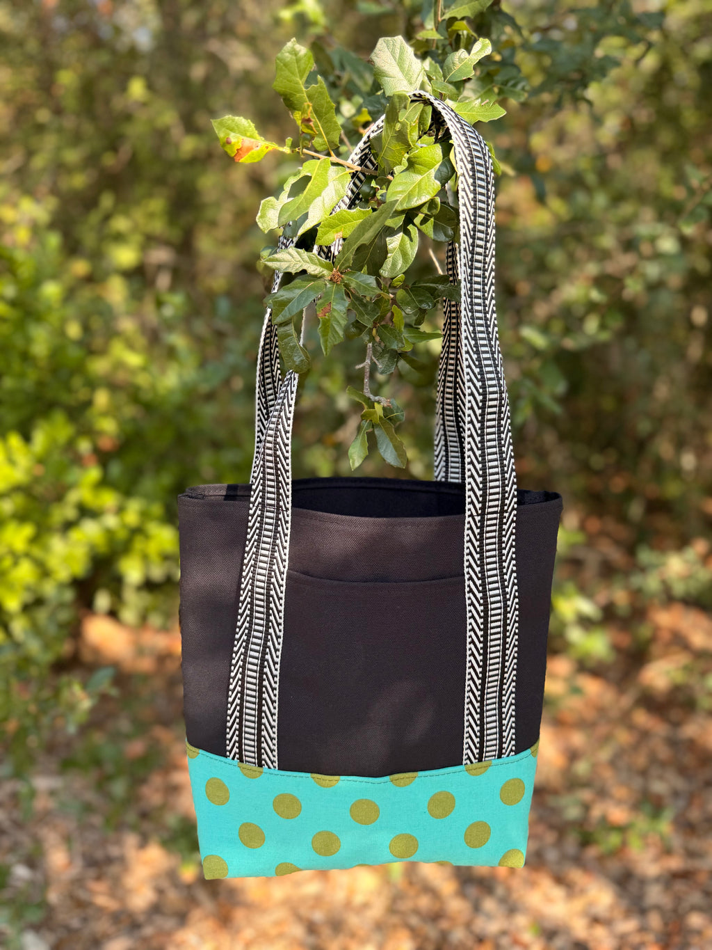 A fabric tote bag  featuring a black upper section, black-and-white patterned handles, and a turquoise bottom panel with green polka dots.