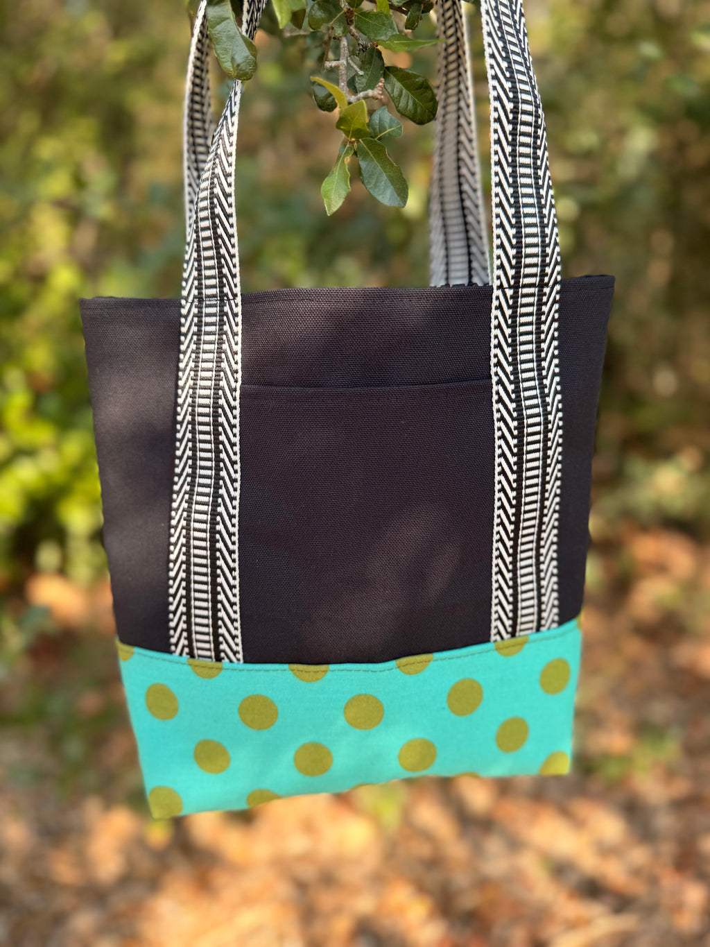 A fabric tote bag  featuring a black upper section, black-and-white patterned handles, and a turquoise bottom panel with green polka dots.