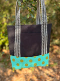 A fabric tote bag  featuring a black upper section, black-and-white patterned handles, and a turquoise bottom panel with green polka dots.