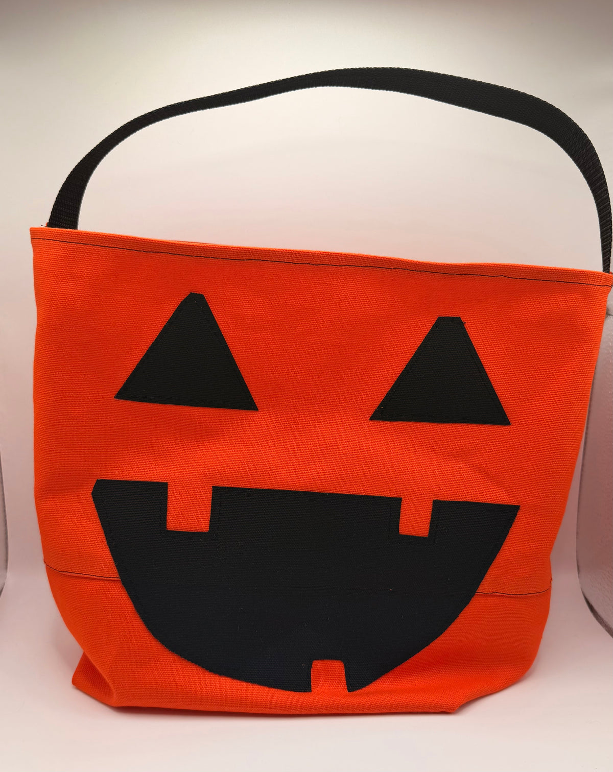 An orange fabric trick-or-treat bucket bag with a black handle and a black jack-o'-lantern face cutout, shown against a plain light background.